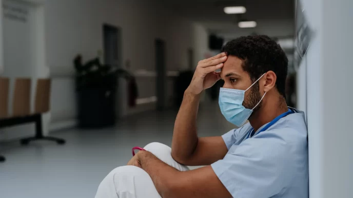 Trabajador migrante con mascarilla descansando en un pasillo hospitalario.