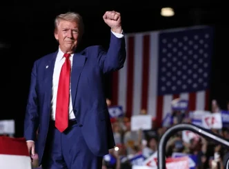 : Donald Trump saludando durante un mitin político ante una bandera de Estados Unidos.