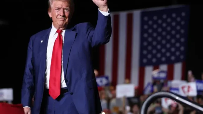 : Donald Trump saludando durante un mitin político ante una bandera de Estados Unidos.