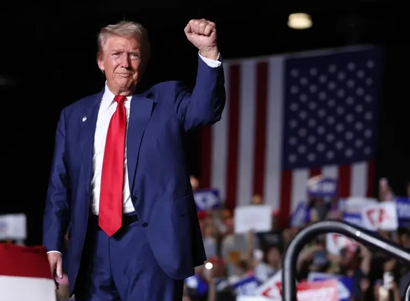 Donald Trump saludando durante un mitin político ante una bandera de Estados Unidos.