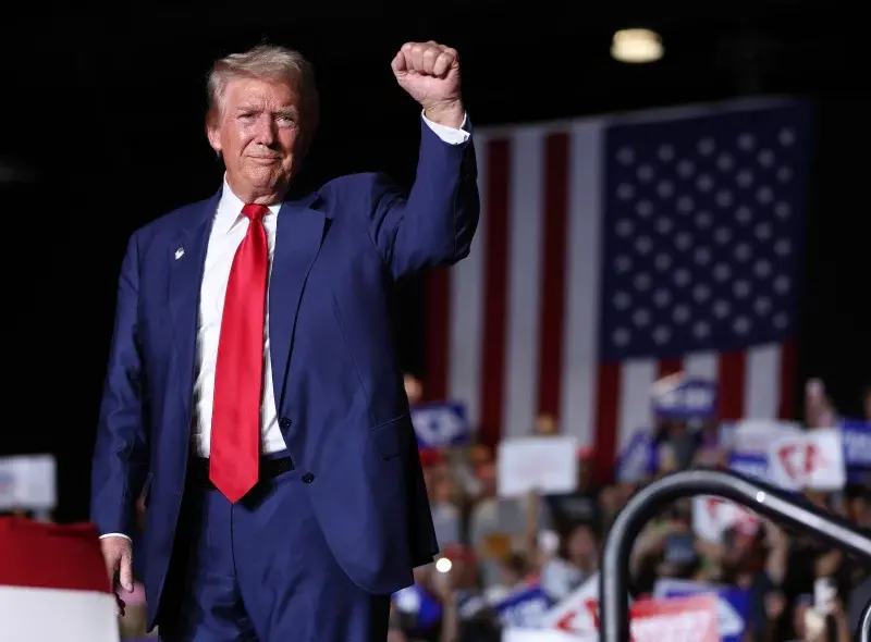 : Donald Trump saludando durante un mitin político ante una bandera de Estados Unidos.