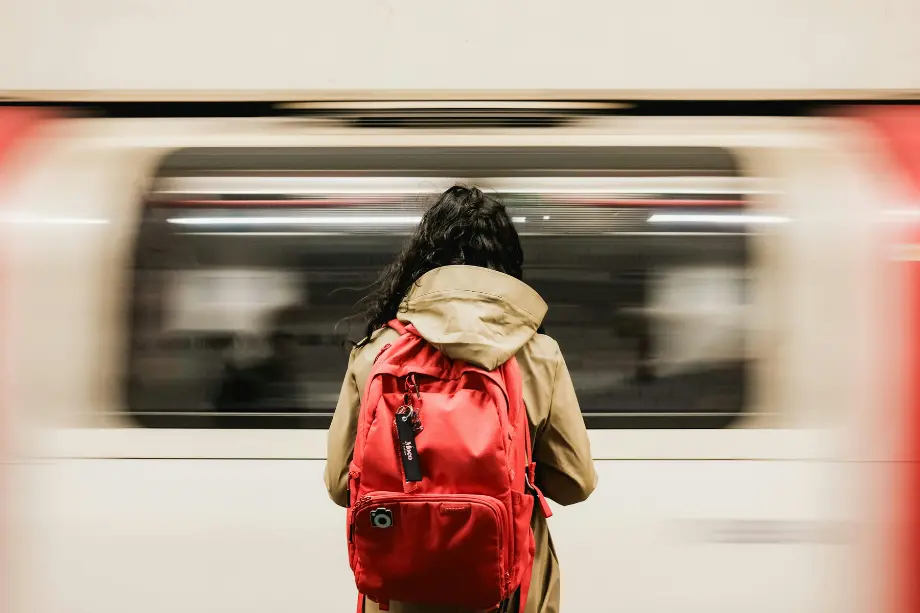 Una joven con mochila roja espera en un andén mientras un tren del metro de Londres pasa rápidamente de fondo.
