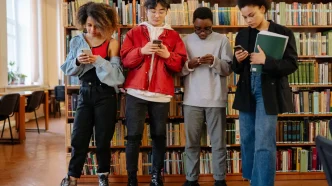 Cuatro jóvenes mirando sus teléfonos móviles en una biblioteca.