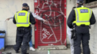 Dos policías de patrulla observan la escena del crimen tras el tiroteo de Keanu Harker. Se aprecian grafitis en la persiana roja del fondo.
