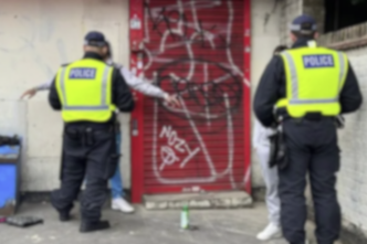 Dos policías de patrulla observan la escena del crimen tras el tiroteo de Keanu Harker. Se aprecian grafitis en la persiana roja del fondo.