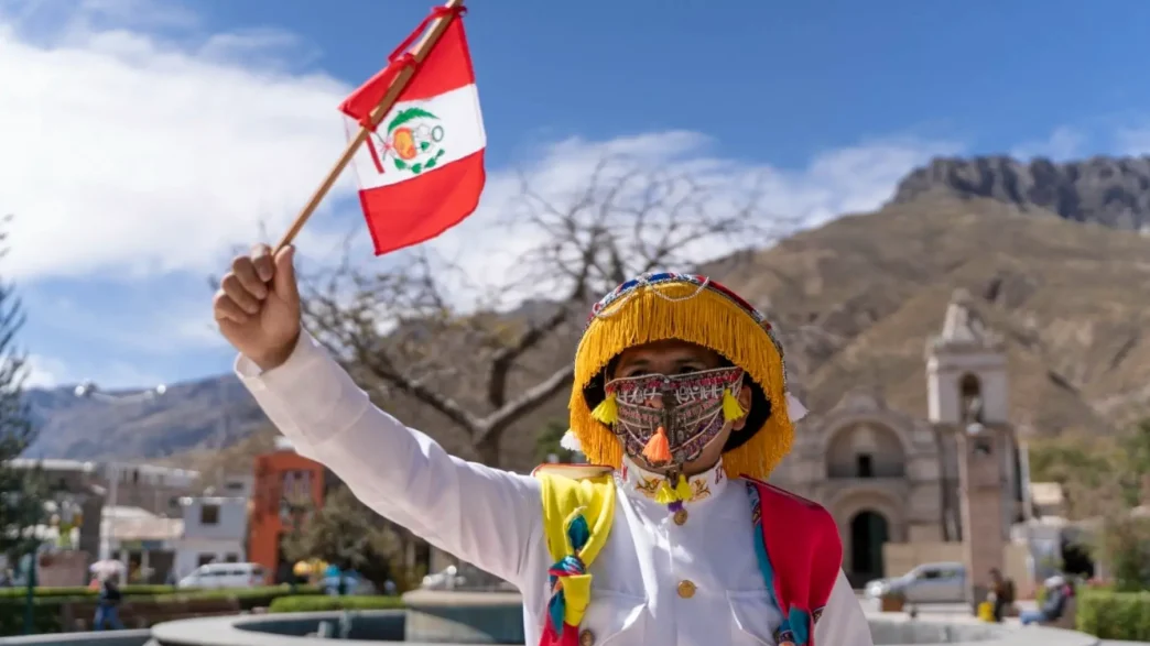 Persona con indumentaria tradicional y máscara sosteniendo una pequeña bandera de Perú durante las celebraciones.