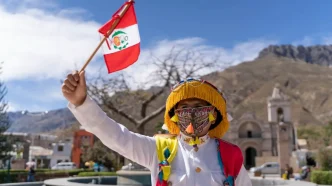 Persona con indumentaria tradicional y máscara sosteniendo una pequeña bandera de Perú durante las celebraciones.
