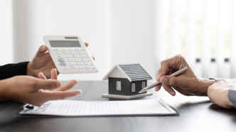 Dos personas analizando un contrato inmobiliario junto a una calculadora y una figura de una casa en miniatura.