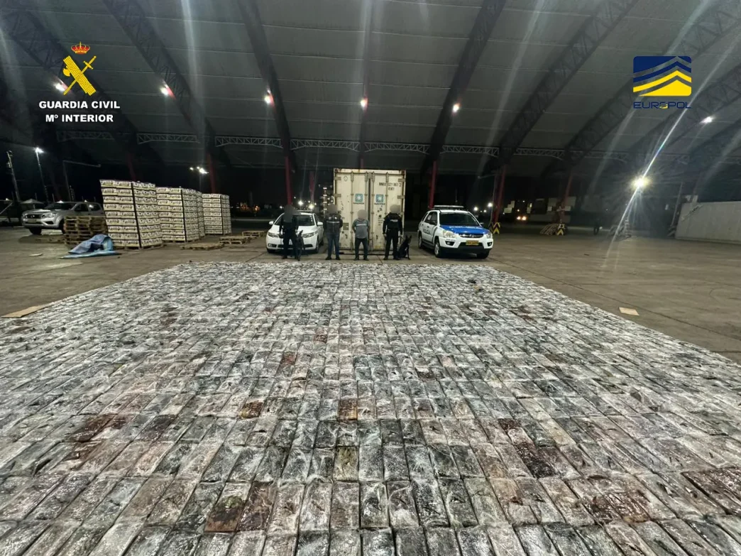 Gran cantidad de paquetes de cocaína incautados en una operación policial junto a un contenedor y vehículos de las autoridades.