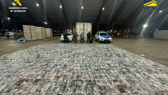 Gran cantidad de paquetes de cocaína incautados en una operación policial junto a un contenedor y vehículos de las autoridades.