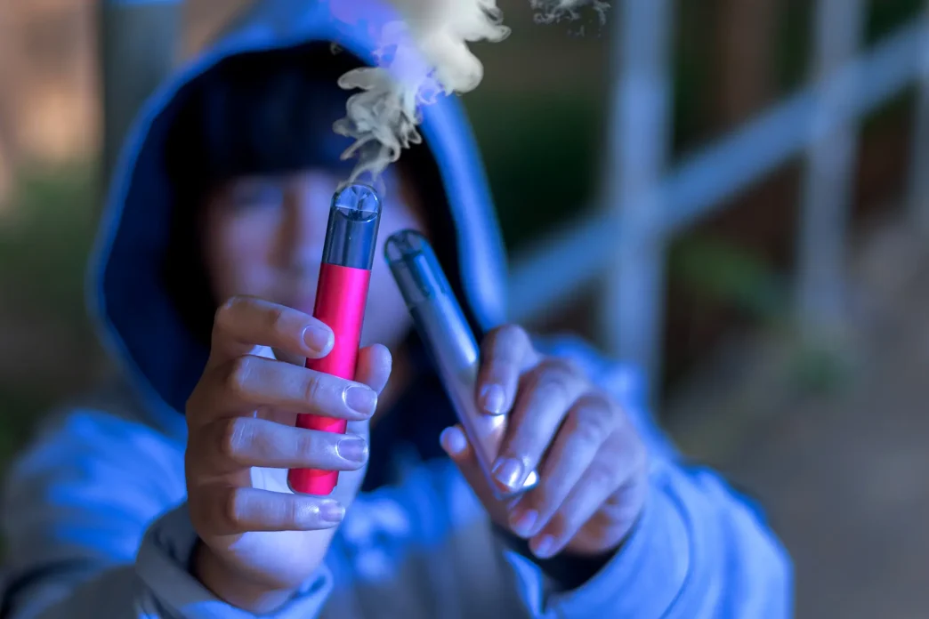 Persona con capucha sosteniendo dos cigarrillos electrónicos en las manos, uno de ellos emitiendo humo.