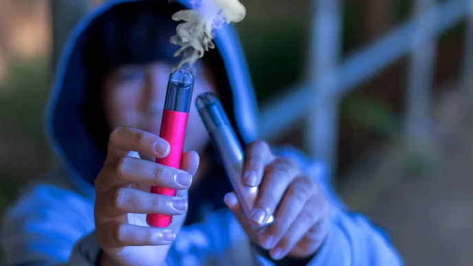 Persona con capucha sosteniendo dos cigarrillos electrónicos en las manos, uno de ellos emitiendo humo.