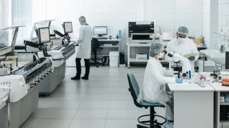 Investigadores con mascarilla y bata de laboratorio analizando muestras de sangre en microscopios y equipos de laboratorio.