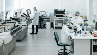 Investigadores con mascarilla y bata de laboratorio analizando muestras de sangre en microscopios y equipos de laboratorio.