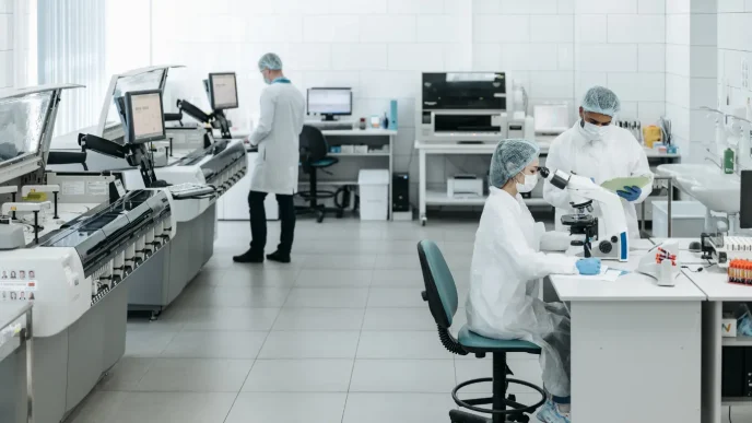 Investigadores con mascarilla y bata de laboratorio analizando muestras de sangre en microscopios y equipos de laboratorio.