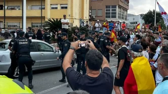 Agentes antidisturbios de la Policía Nacional rodeando un coche destrozado entre una multitud de manifestantes en Ceuta.