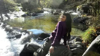 La joven estudiante española Claudia Tacoronte Aguilera sonriendo sentada en una roca junto a un río en la naturaleza.