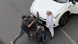 Un hombre sujetando brutalmente por el cuello a un ciclista frente a un automóvil blanco en la vía pública.