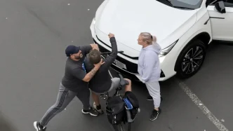 Un hombre sujetando brutalmente por el cuello a un ciclista frente a un automóvil blanco en la vía pública.