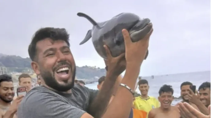 Hombre exhibiendo fuera del agua a una cría de delfín en la playa Trampolín de Ceuta rodeado de personas.