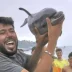 Hombre exhibiendo fuera del agua a una cría de delfín en la playa Trampolín de Ceuta rodeado de personas.