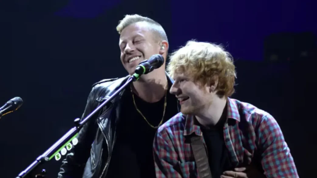 Ed Sheeran tocando la guitarra junto al rapero Macklemore sonriendo en un escenario.