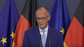 El canciller alemán Friedrich Merz con traje azul y gafas comparece en atril ante las banderas de Alemania y la Unión Europea.