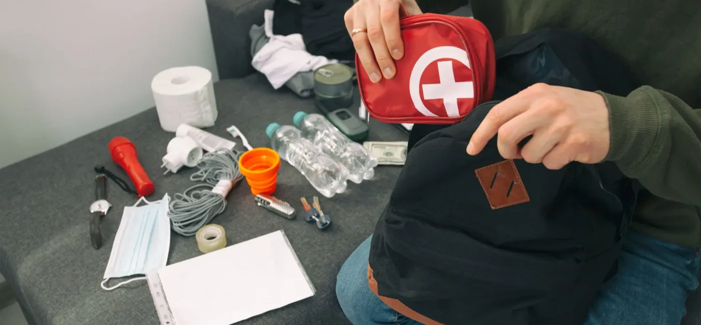 Persona guardando un botiquín de primeros auxilios rojo en una mochila de emergencia junto a botellas de agua, linterna y alimentos.