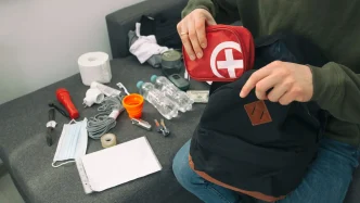 Persona guardando un botiquín de primeros auxilios rojo en una mochila de emergencia junto a botellas de agua, linterna y alimentos.