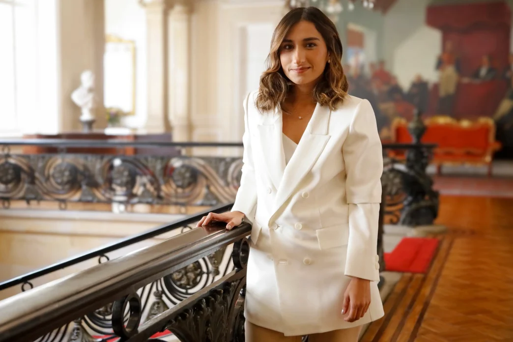 Laura Sarabia vestida con un traje blanco posando junto a una barandilla en un edificio institucional.