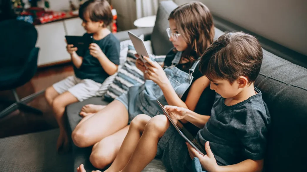 Niños y adolescentes sentados en un sofá utilizando tabletas y teléfonos móviles.
