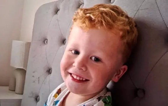 Retrato del niño Noah Woods sonriendo a la cámara apoyado en un respaldo gris.