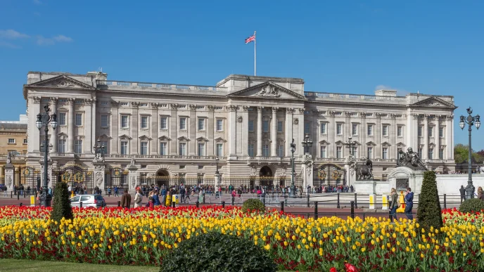 Fachada principal del Palacio de Buckingham con flores en los jardines y la bandera británica ondeando en el tejado