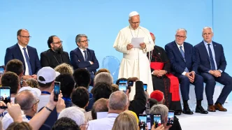 El papa León XIV leyendo un discurso en el escenario rodeado de autoridades religiosas y civiles ante un público que toma fotografías con teléfonos móviles.