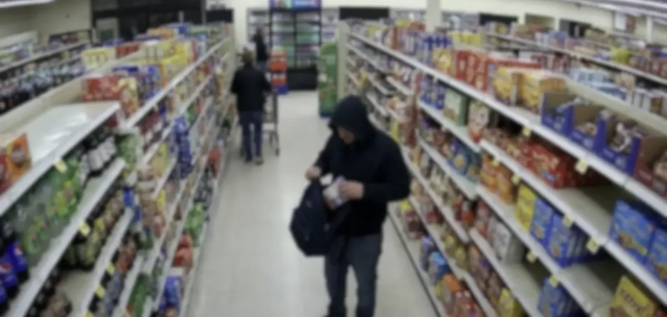 ombre encapuchado guardando productos en un bolso dentro del pasillo de un supermercado captado por cámara de seguridad.