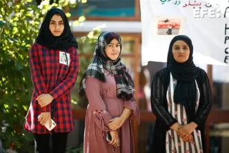 Tres mujeres afganas vestidas con hiyab y túnicas tradicionales posan de pie en un espacio público.