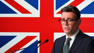 El primer ministro Andy Burnham hablando en un atril ante la bandera del Reino Unido.