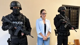 Dos agentes de la policía táctica con pasamontañas negros y fusiles de asalto flanquean a una mujer con gafas en el interior de una sala.