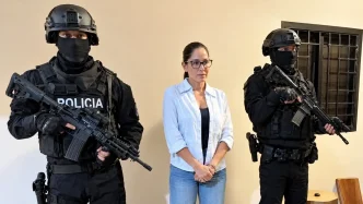 Dos agentes de la policía táctica con pasamontañas negros y fusiles de asalto flanquean a una mujer con gafas en el interior de una sala.