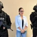 Dos agentes de la policía táctica con pasamontañas negros y fusiles de asalto flanquean a una mujer con gafas en el interior de una sala.