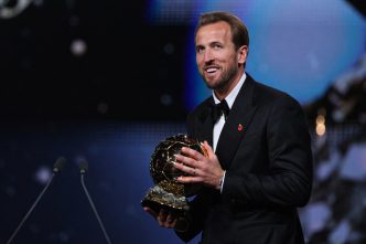 Harry Kane sonriendo en el escenario mientras sostiene el trofeo del Balón de Oro 2026 vistiendo un esmoquin negro.