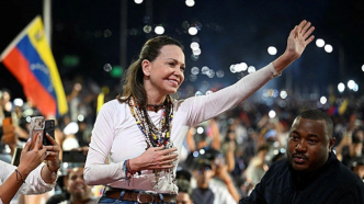 María Corina Machado saluda sonriente con la mano levantada entre una multitud de seguidores y banderas de Venezuela.