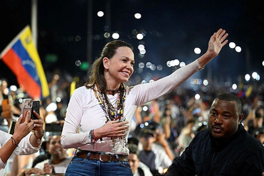 María Corina Machado saluda sonriente con la mano levantada entre una multitud de seguidores y banderas de Venezuela.