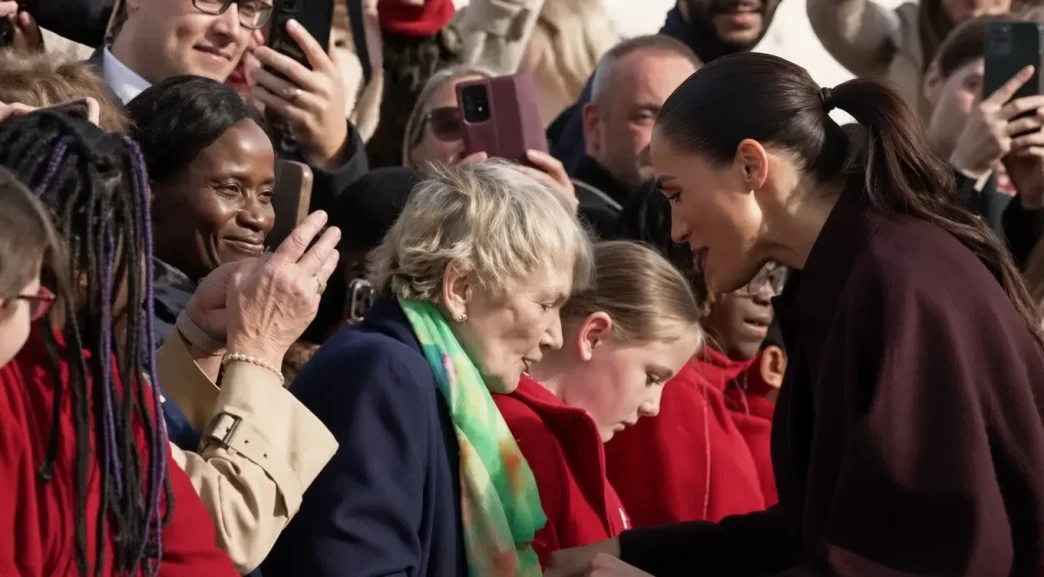 Meghan Markle con abrigo burdeos inclinándose sonriente para saludar a la multitud.
