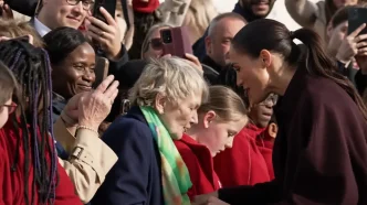 Meghan Markle con abrigo burdeos inclinándose sonriente para saludar a la multitud.