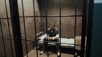Un hombre sentado en la cama de una celda de prisión tras unos barrotes de hierro.