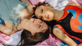 Dos personas jóvenes recostadas sobre una bandera del orgullo transgénero de colores rosa, blanco y azul.