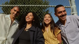Cuatro jóvenes latinos sonrientes al aire libre junto a una valla metálica de protección.