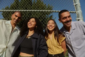 Cuatro jóvenes latinos sonrientes al aire libre junto a una valla metálica de protección.
