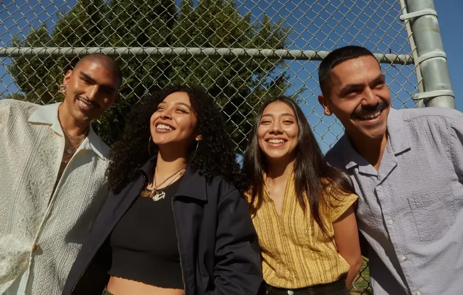 Cuatro jóvenes latinos sonrientes al aire libre junto a una valla metálica de protección.